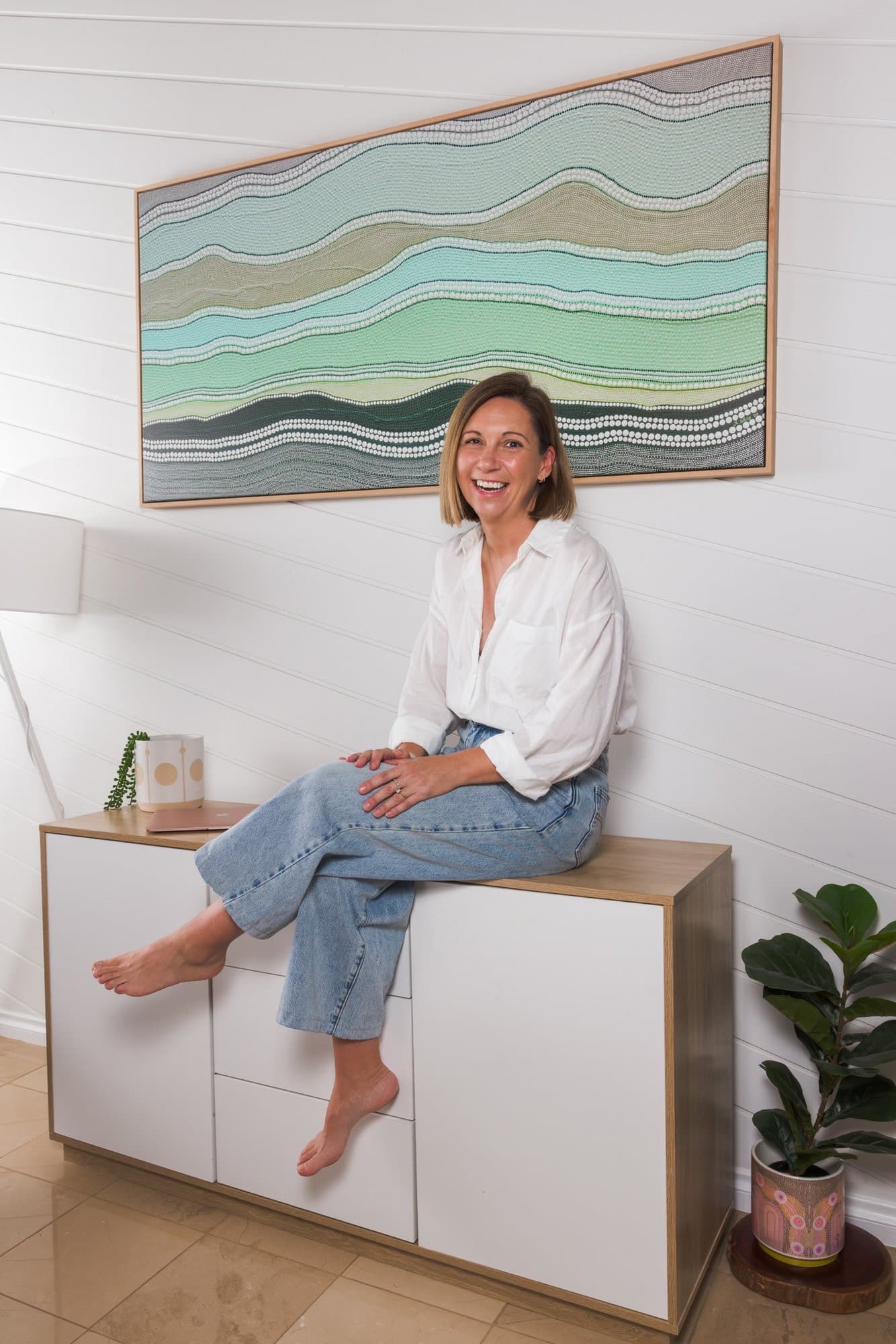 Steph sitting on a sideboard, laughing at the camera, a green dot-art painting on the wall behind her