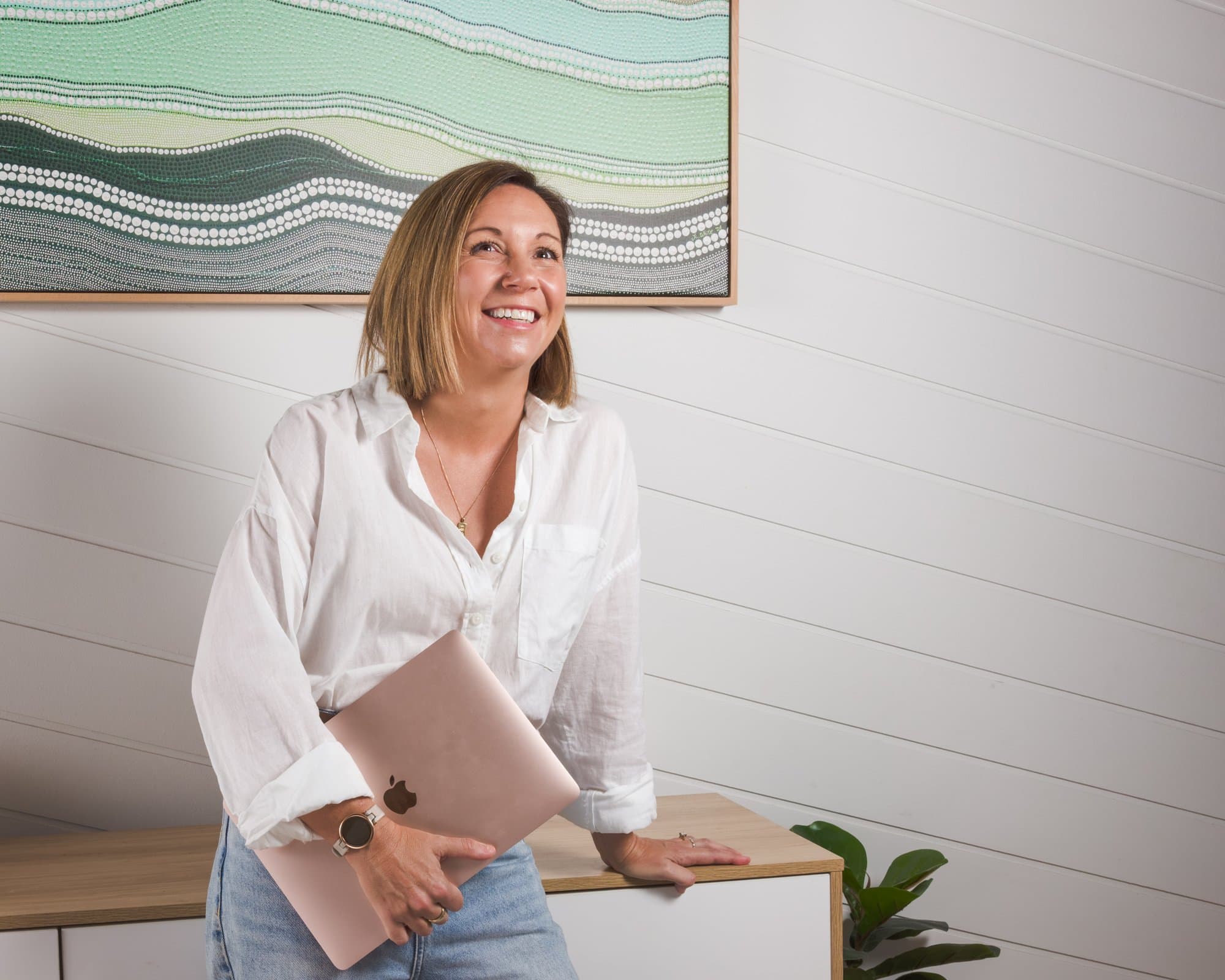 Steph leaning on a sideboard with her laptop under one arm, smiling