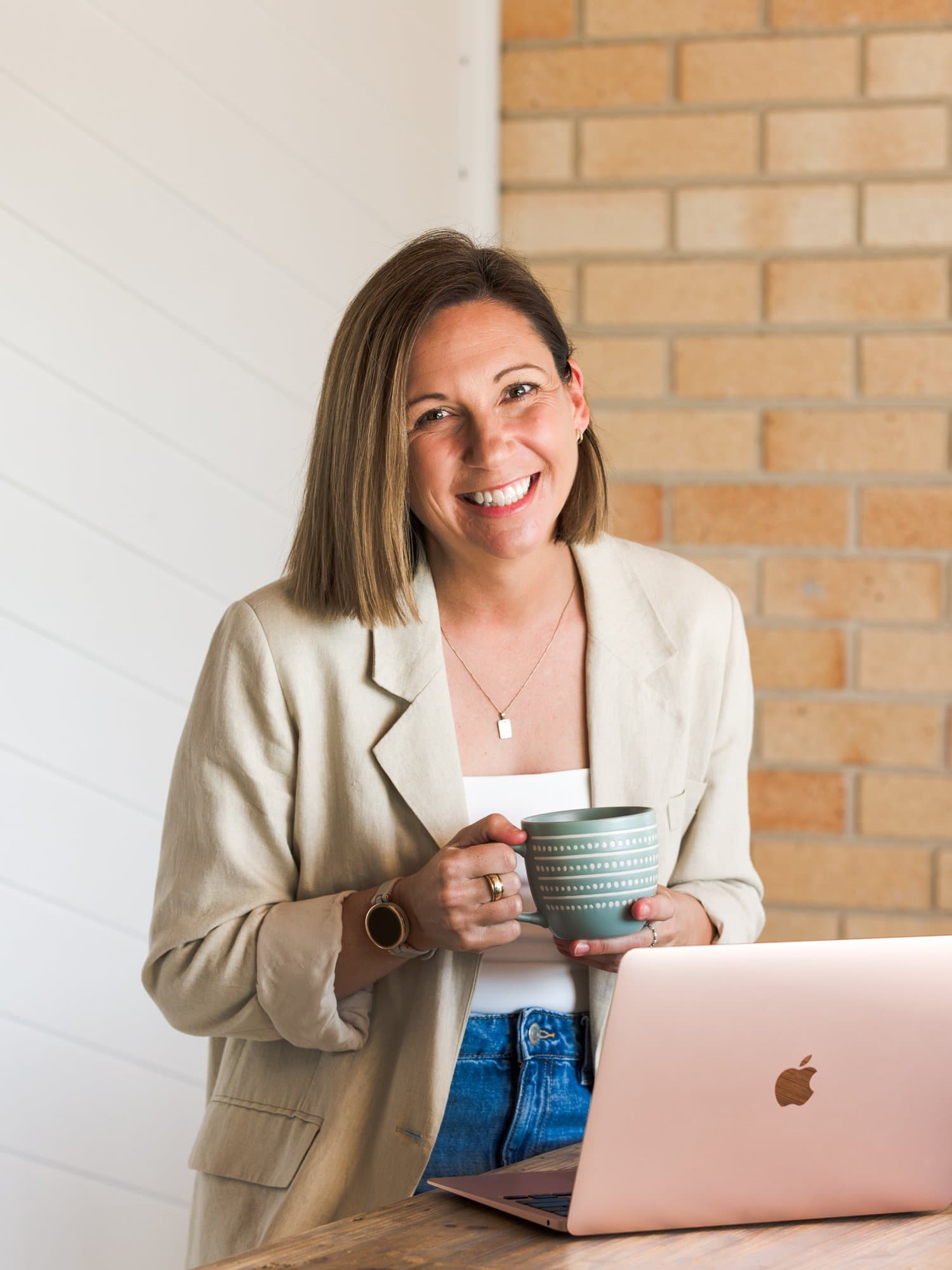 Steph in a linen blazer holding a mug, laptop open in front of her