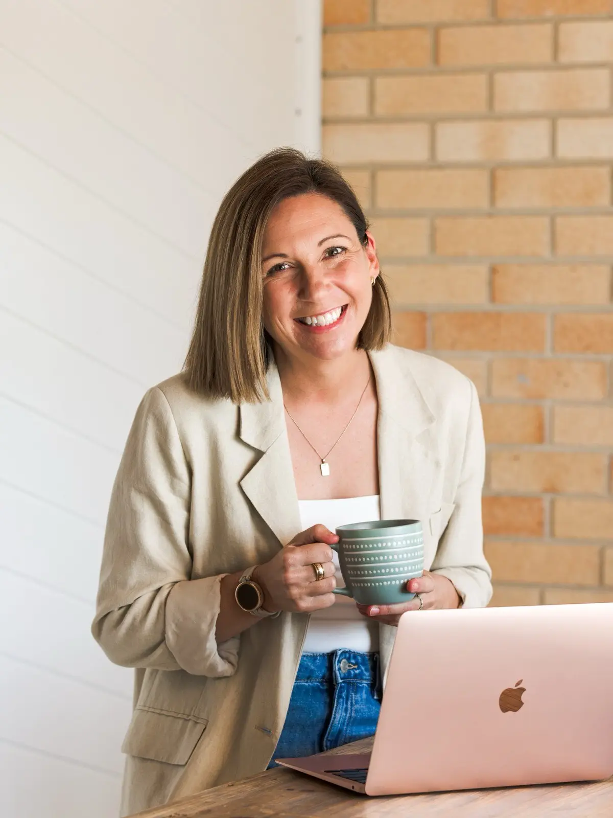 Steph smiling at the camera holding a mug beside her laptop